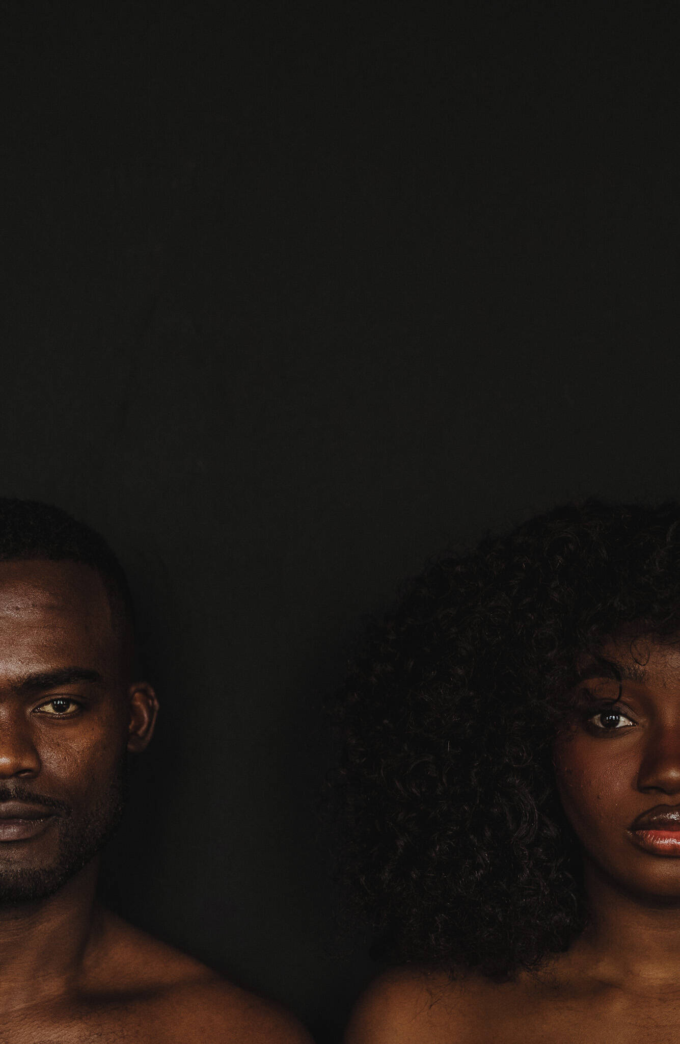Photographic portrait with a dark background. Two faces, both in the bottom corners of the image, one male and one female.